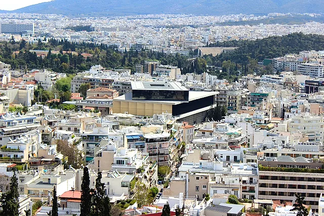 The Acropolis Museum, a modern building of glass and pale stone among the houses of Athens, seen from a hill