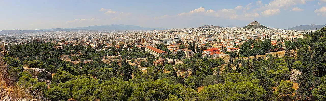 Wide panorama of Athens seen from the Acropolis hill, with pine trees in the foreground and the city stretching to the hills on the horizon
