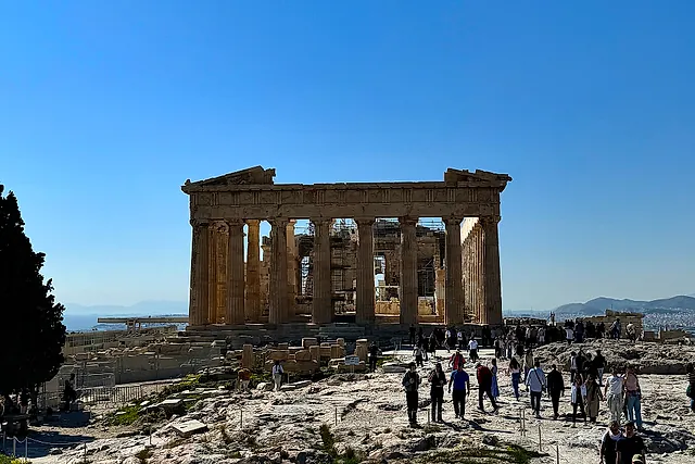 The Parthenon seen across the rocky summit of the Acropolis, with visitors walking in front and a clear blue sky